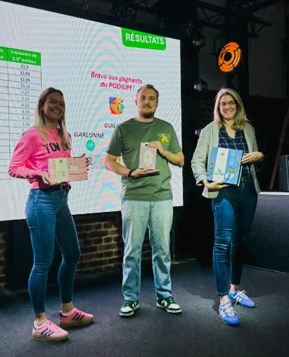 Trois personnes debout sur une scène, chacune tenant un coffret, devant un écran affichant « RÉSULTATS » et « Bravo aux gagnants du PODIUM ! ».